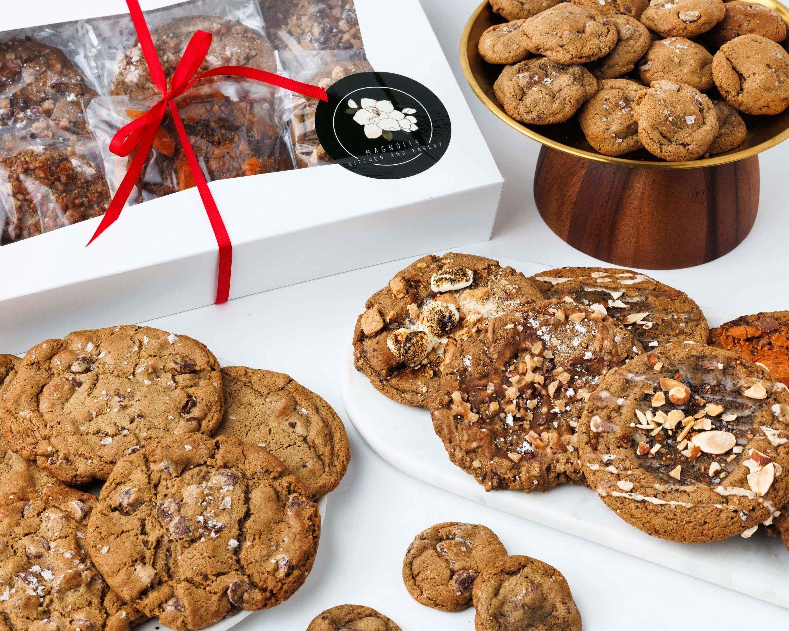Magnolia corporate cookie gift box tied with a red ribbon beside plates of chef crafted cookies and mini chocolate chip cookies