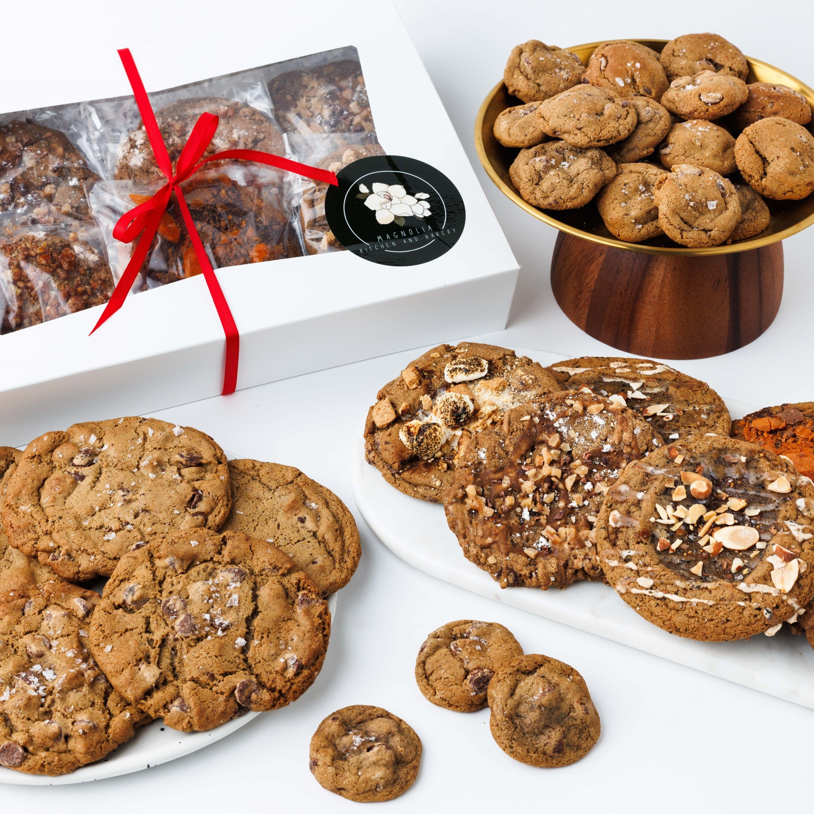 Magnolia corporate cookie gift box tied with a red ribbon beside plates of chef crafted cookies and mini chocolate chip cookies