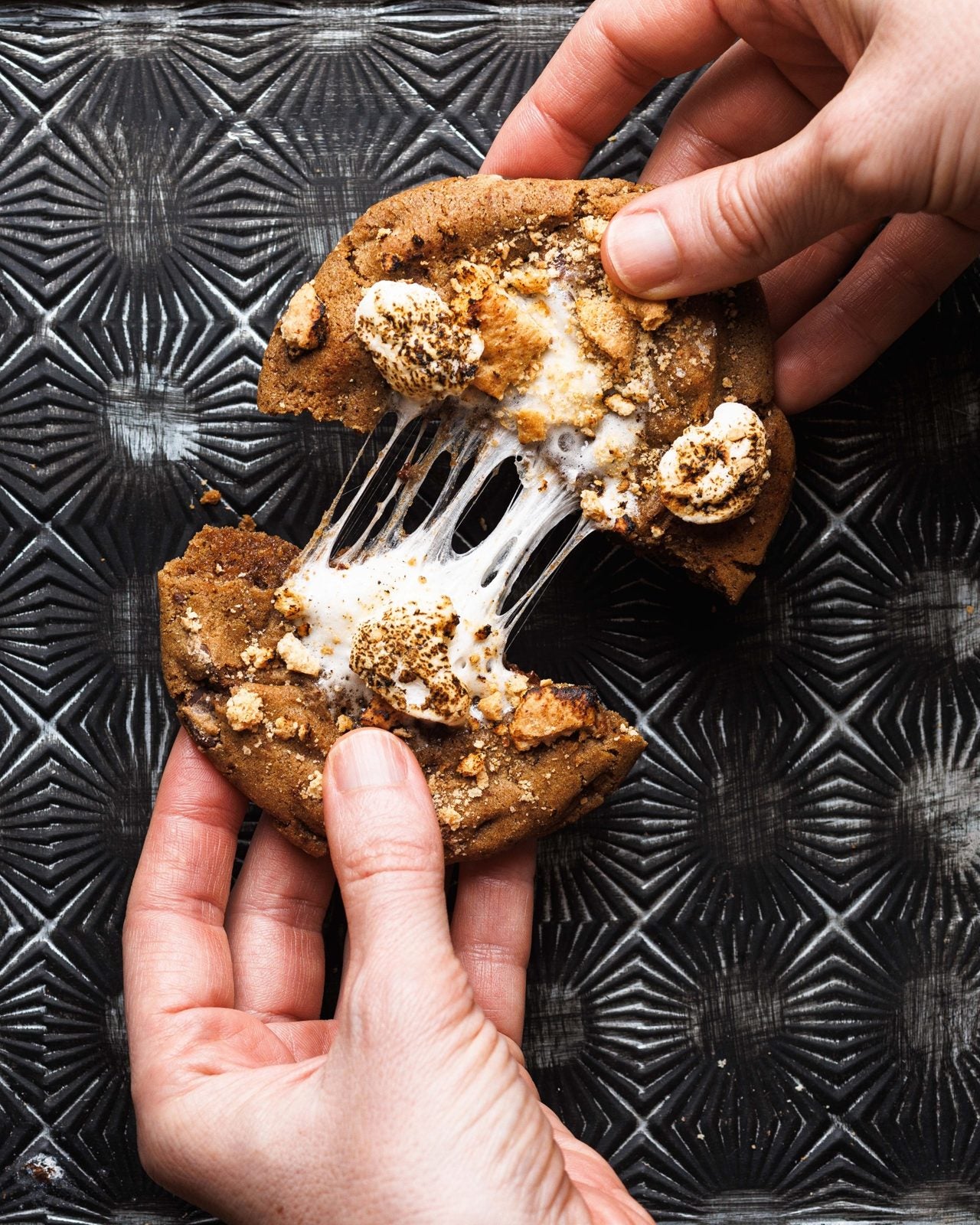 Hands pulling apart a s'mores cookie with stretchy fire-roasted marshmallow and graham crumbs